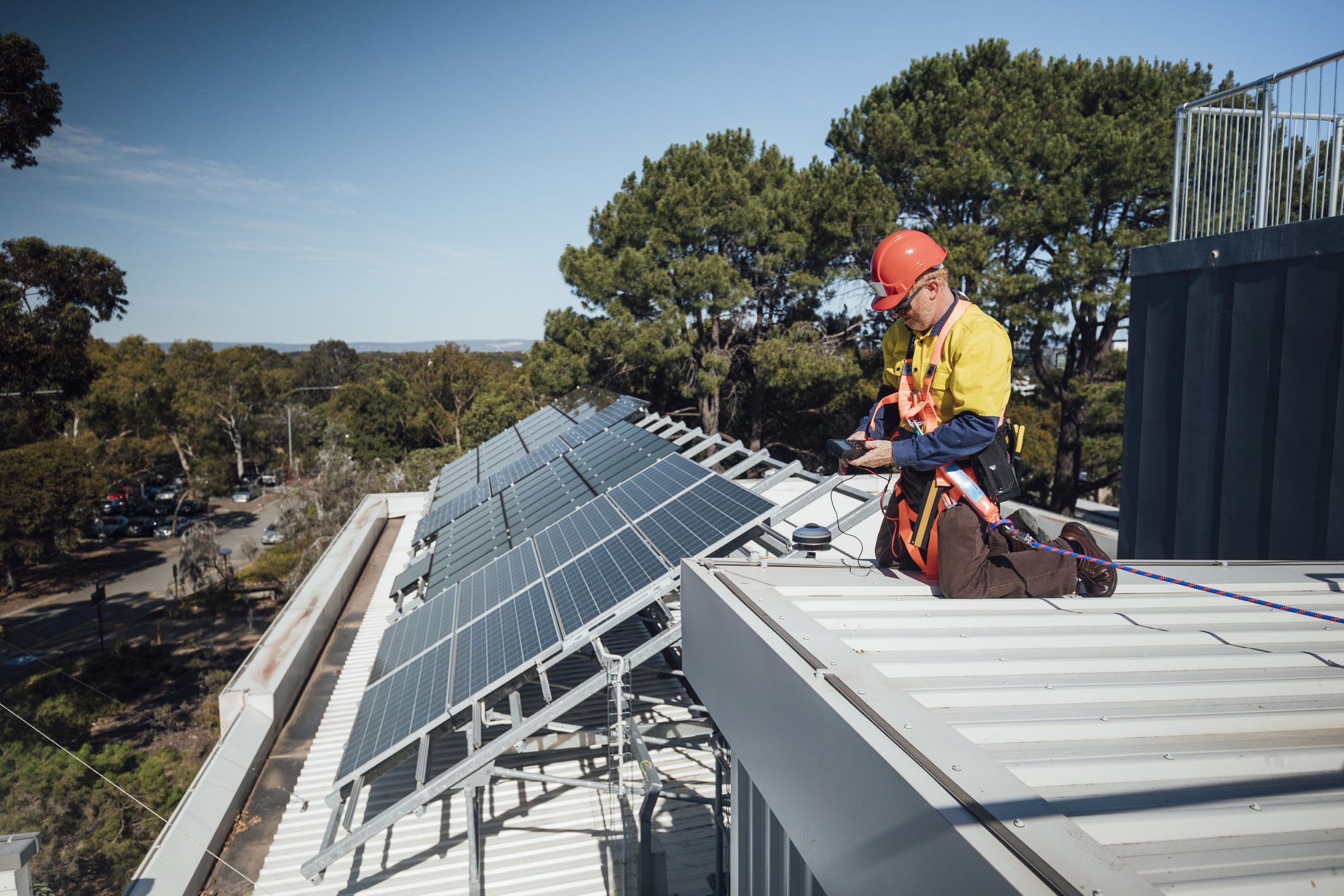 A mature man at work, repairing some solar cell panels on the roof a university. Wearing his overalls, harness and hardhat.